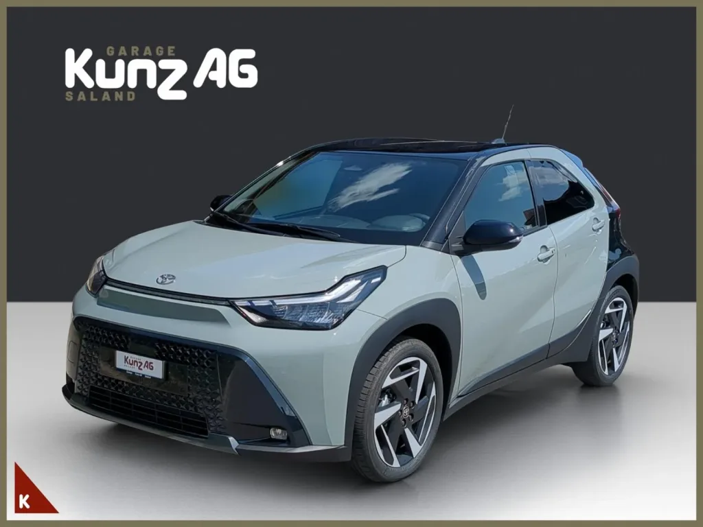 Schräge Frontansicht eines hellgrauen Toyota Aygo X Style mit schwarzem Dach vor grauem Hintergrund.